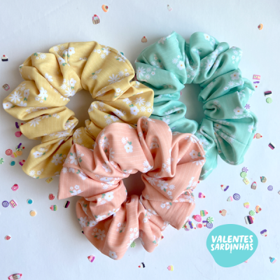 Três scrunchies florais amarelo, verde água e rosa sobre fundo branco com confetes coloridos