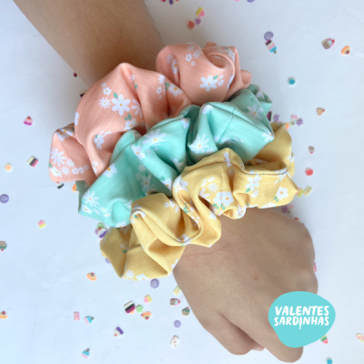 Três scrunchies florais coloridos usados no pulso sobre fundo branco com confetis.
