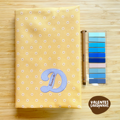 Capa amarela com margaridas e letra D azul claro, caneta preta e palete de cores azuis num fundo de madeira