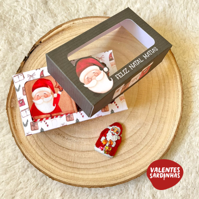 Caixa de presente de Natal com figura do Pai Natal e texto, junto de cartão e chocolate, sobre base de madeira redonda.