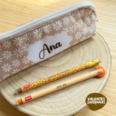 Estojo bege com flores brancas e o nome Ana, dois lápis com tema girafa sobre base de madeira
