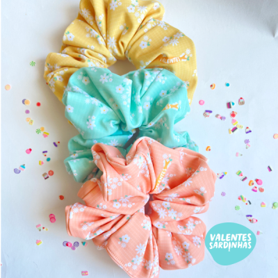 Três scrunchies florais amarelo, verde água e coral sobre fundo branco com confetes.