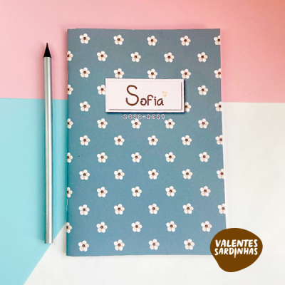Caderno azul com flores brancas e etiqueta com texto 'Sofia 2023-2024' ao lado de caneta