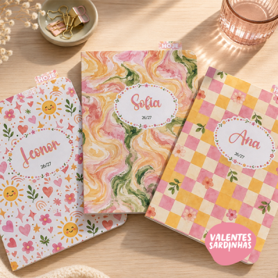 Três cadernos coloridos com padrões florais e geométricos e nomes nas capas, sobre mesa clara com clips e copo rosa