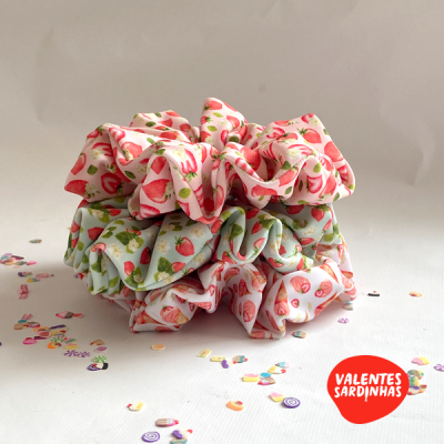 Três scrunchies com padrão de morangos empilhados sobre superfície branca com confetes e logotipo Valentes Sardinhas
