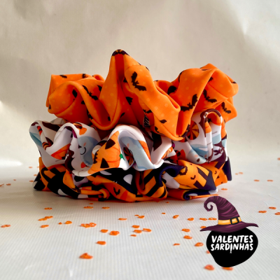 Três scrunchies coloridos com padrões de Halloween sobre superfície branca com confetes laranja