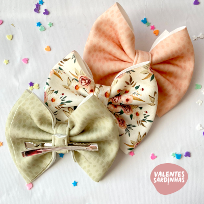 Três laços de cabelo coloridos com padrões e presilha metálica, sobre superfície branca com confetes coloridos.