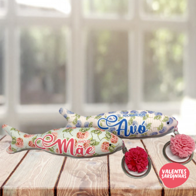 Almofadas em formato de sardinha com padrões florais e textos AMOR Mãe e ADORAMOS Avó sobre base de madeira com flores de tecido e selo VALENTES SARDINHAS.