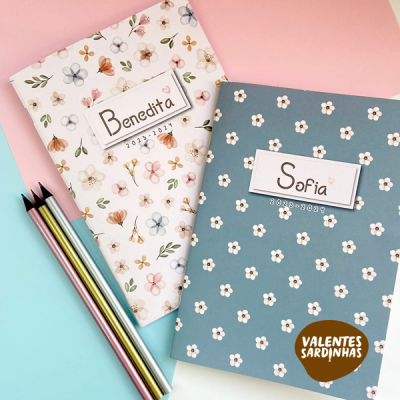 Dois cadernos florais com nomes 'Benedita' e 'Sofia' e data 2023-2024, junto a lápis coloridos sobre fundo azul e rosa
