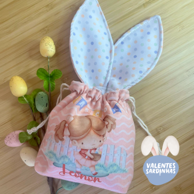 Saco de pano cor-de-rosa com desenho de coelho e orelhas de coelho brancas com bolinhas coloridas, sobre superfície de madeira clara com ovos decorativos e folhas verdes.