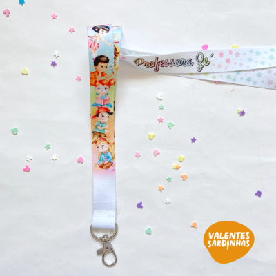 Lanyard branco com desenhos de crianças e texto 'Professora Gé' com gancho metálico, sobre superfície branca com confetis coloridos.