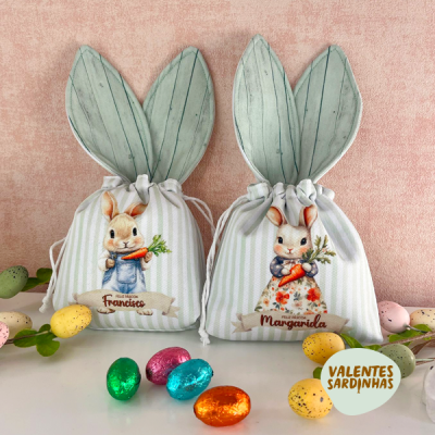 Duas bolsas em tecido com orelhas de coelho e ilustrações de coelhos, com ovos de chocolate coloridos à frente, fundo rosa claro.