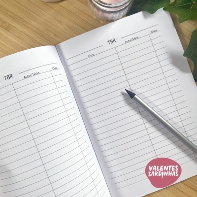 Cadernos abertos com colunas para TBR contendo caneta sobre página em mesa de madeira clara e folhas verdes ao lado