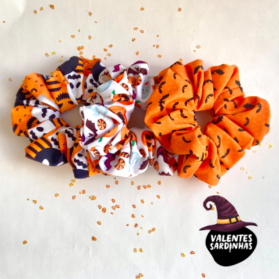 Duas scrunchies de tecido com padrões de Halloween em fundo branco e logo Valentes Sardinhas