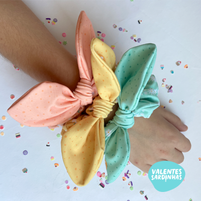 Três pulseiras de tecido com laços grandes em tons pastel no pulso.