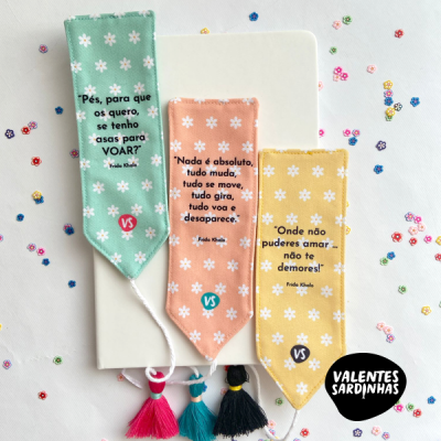 Três marcadores de livro de tecido com flores brancas e frases de Frida Kahlo em português, com tassel, sobre fundo branco com lantejoulas.