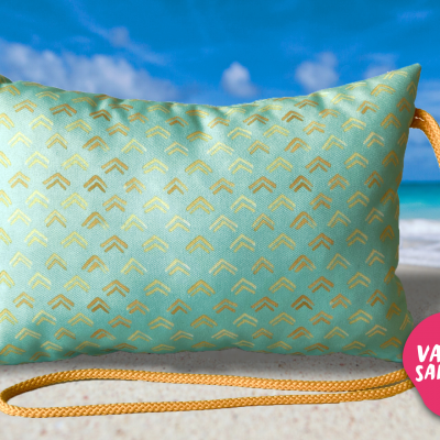 Almofada azul com padrão dourado, cordão laranja e etiqueta rosa, na praia