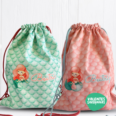 Dois sacos de cordão com padrão de escamas e ilustração de sereia, em verde e rosa, nome 'Beatriz' e logotipo 'VALENTES SARDINHAS'