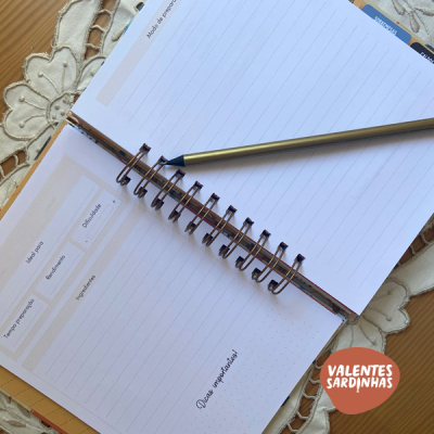 Caderno aberto com caneta dourada sobre páginas com linhas azuis e toalha bordada