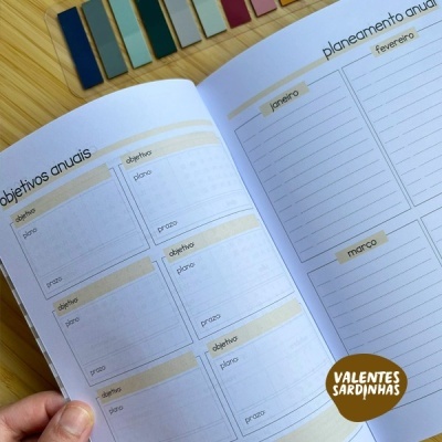 Planner aberto com seções para objetivos anuais e planeamento anual, suportado por uma mão, com lápis coloridos ao fundo