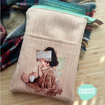 Necessaire rosa com estampa de mulher a ler livro e fecho verde, ao lado de manta xadrez e superfície em madeira clara