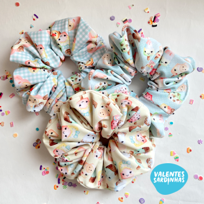 Três scrunchies com padrões de gatinhos em tons pastel sobre fundo branco com confetis coloridos
