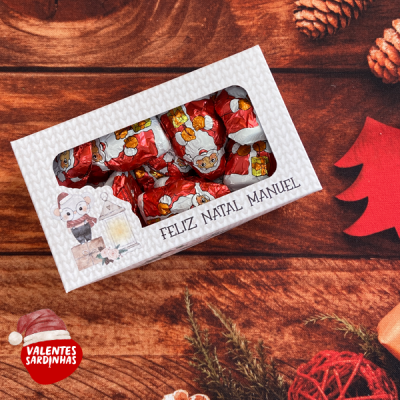 Caixa branca com chocolates embrulhados decorados com Pai Natal, mensagem 'FELIZ NATAL MANUEL' e decoração natalícia de fundo