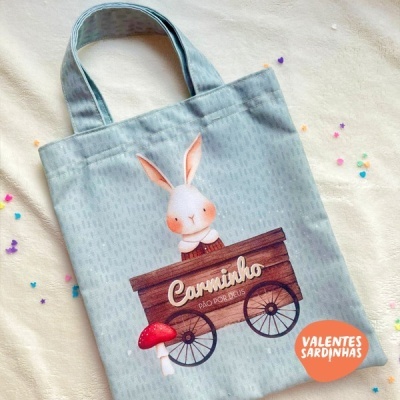 Saco de tecido azul claro com ilustração de coelho e caixa de madeira com texto 'Caminho PÃO POR DEUS'