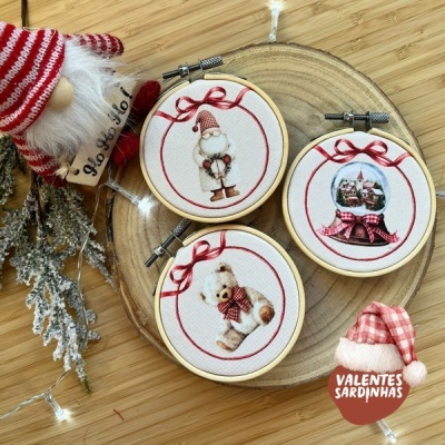 Três bordados de Natal em molduras redondas com desenhos festivos colocados sobre madeira clara ao lado de boneco natalício.