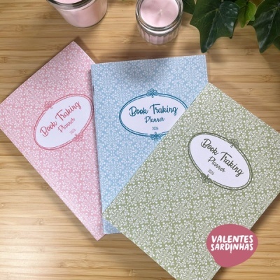 Três cadernos coloridos Book Tracking Planner 2026 com velas e folhas verdes sobre mesa de madeira