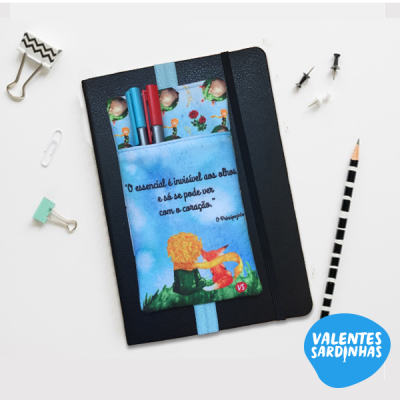 Caderno preto com bolso azul ilustrado e frase em português, canetas dentro, sobre mesa branca com acessórios de papelaria.