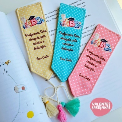Marcadores de livros de tecido amarelo, azul e rosa com mensagem para professora Sara e franjas coloridas.