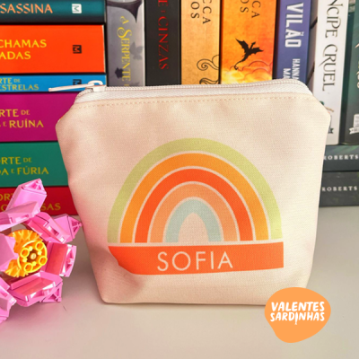 Bolsa de tecido bege com arco-íris colorido e nome Sofia, sobre uma mesa branca com livros e uma flor rosa ao lado