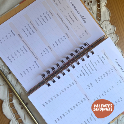 Caderno espiral aberto com tabelas de conversões e medidas sobre superfície de madeira clara com pano bordado