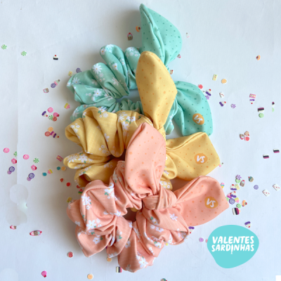 Três scrunchies coloridos com orelhas de coelho e padrão floral sobre fundo branco com confetes coloridos