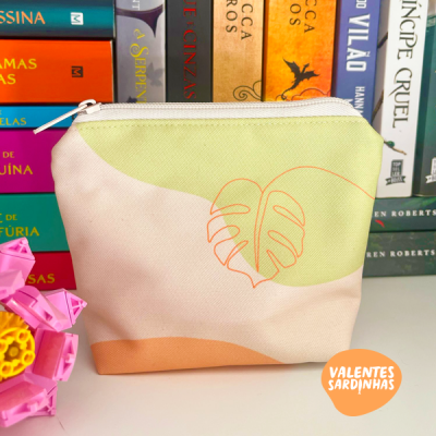 Necessaire de tecido com padrão verde claro, bege, castanho e folha laranja, com texto VALENTES SARDINHAS em embalagem laranja, em mesa branca com livros atrás