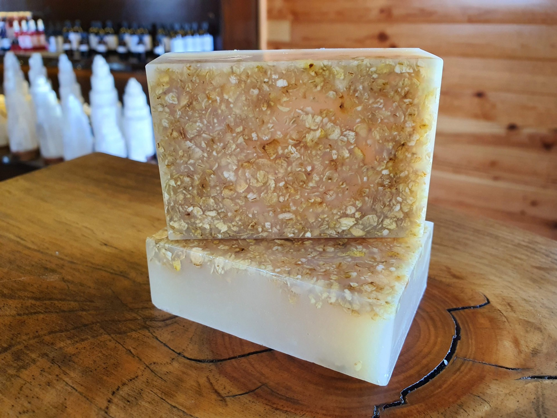 Sabonete Esfoliante Leite de Cabra e Aveia Dois sabonetes artesanais retangulares com flocos cor de bege claro na parte superior, sobre superfície de madeira