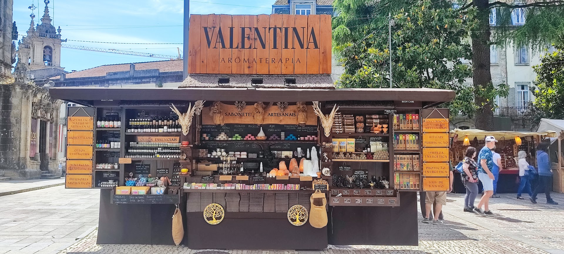 Banca de madeira ao ar livre com produtos artesanais e cartaz VALENTINA A LOJA DA PRAÇA