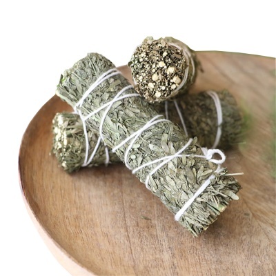 Varas de folhas secas verdes amarradas com fio branco em superfície de madeira redonda com tigela de ervas secas ao fundo