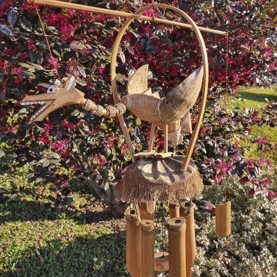 Sino dos ventos de madeira em forma de ave pendurado em jardim com arbustos e flores vermelhas