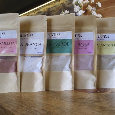 Cinco embalagens pequenas com pós coloridos da marca Valentina Body Care alinhadas sobre uma mesa de madeira.