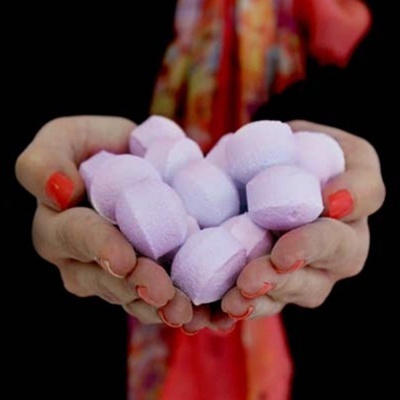 Mãos segurando pastilhas lilás em fundo preto com detalhes vermelhos e laranja