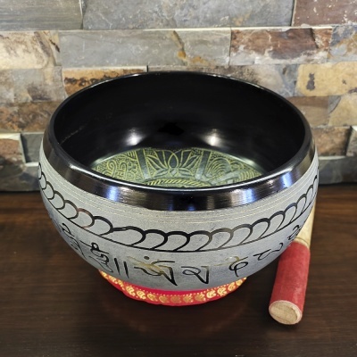 Taça tibetana prateada com gravações pretas, interior preto com padrão verde, sobre almofada vermelha, com rolo vermelho na madeira e fundo em pedra