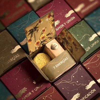 Caixa aberta com esponja e frasco etiquetados 7 ENERGIAS sobre caixas coloridas com textos em espanhol