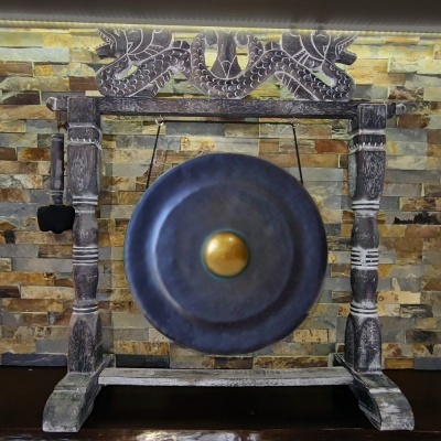 Gongo de metal preto com centro dourado numa estrutura de madeira esculpida, parede de tijolos decorativos