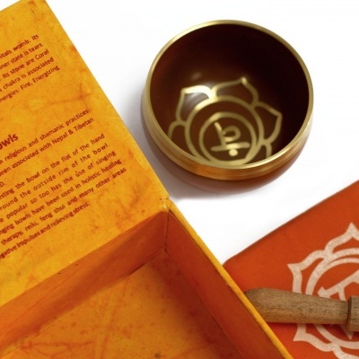 Taça tibetana com símbolo dourado, caixa amarela, tecido laranja com padrão branco e bastão de madeira.