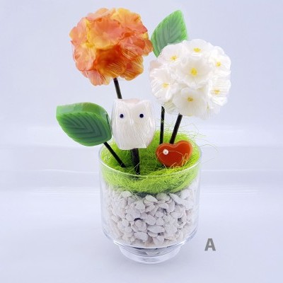 Arranjo decorativo em copo de vidro com flores laranja e branca, folhas verdes, coruja branca e coração vermelho