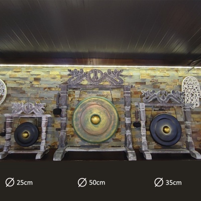 Três gongos decorativos com armações esculpidas em madeira e superfície metálica, exibidos contra parede de pedra.