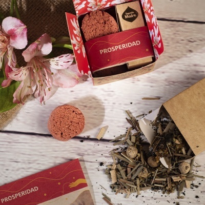 Caixa decorativa com biscoitos e pacote com a palavra PROSPERIDAD, ervas secas e flores cor-de-rosa sobre madeira clara.