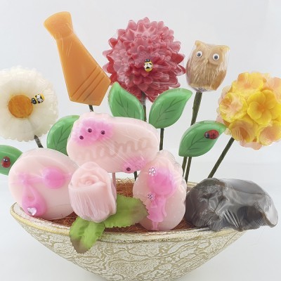 Arranjo decorativo colorido de flores em base oval com folhas verdes e insetos decorativos.